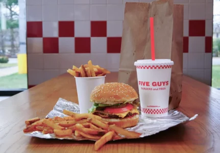 Five Guys burger, fry cup, and drink on a wooden table with red and white checker tiles in background.
