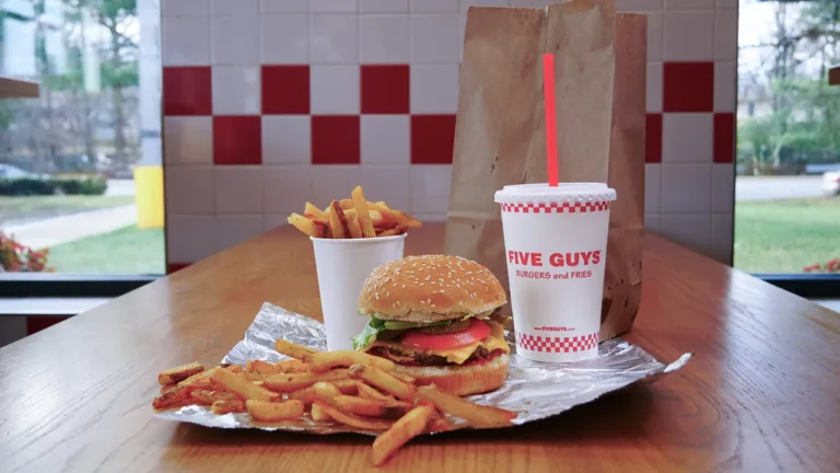 Five Guys burger, fry cup, and drink on a wooden table with red and white checker tiles in background.